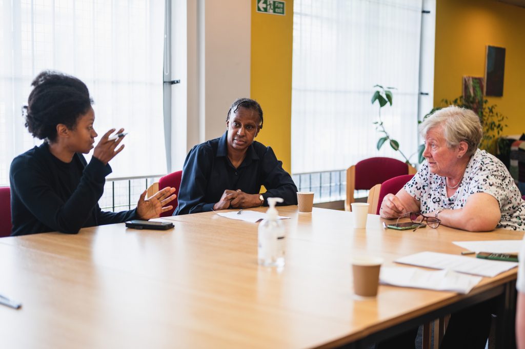 Three people seated around a table in a bright room with large windows, having a conversation.